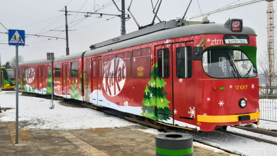 Czerwony tramwaj ozdobiony grafiką choinek i śniegu. Tramwaj stoi na przystanku.