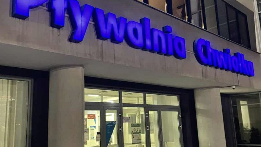 Widok spod wejścia do białego budynku basenu. Nad wejściem niebieski neon z napisem "Pływalnia Chwiałka".