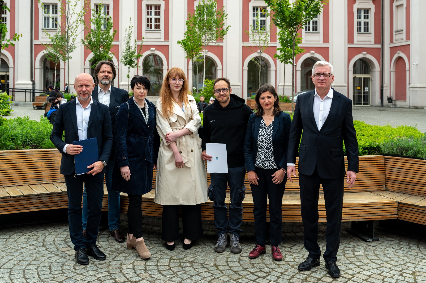 Siedem osób na tle zieleni i biło-różowego budynku