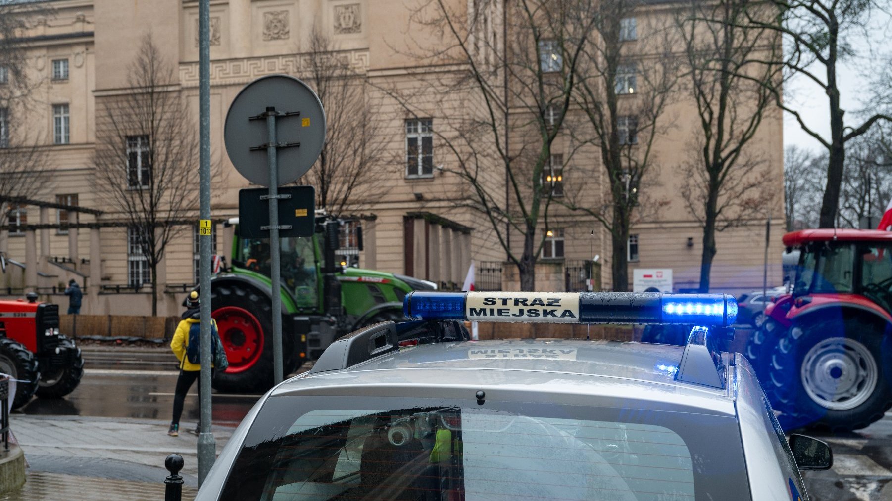 Zdjęcie przedstwia sznur ciągników na ulicy oraz samochód straży miejskiej.