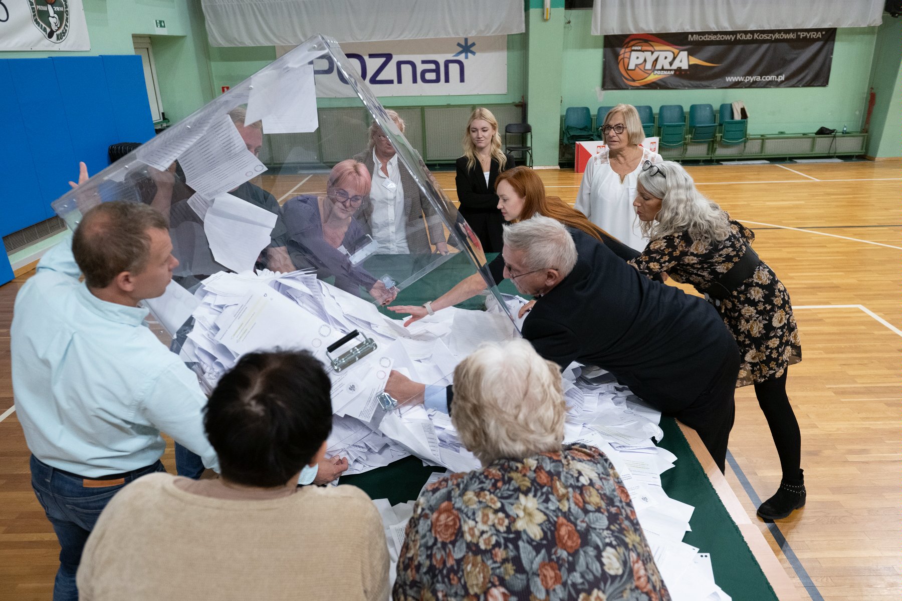 Zdjęcie przedstawia grupę osób, członków OKW, wysypujących z urny wyborczej karty do głosowania. - grafika artykułu