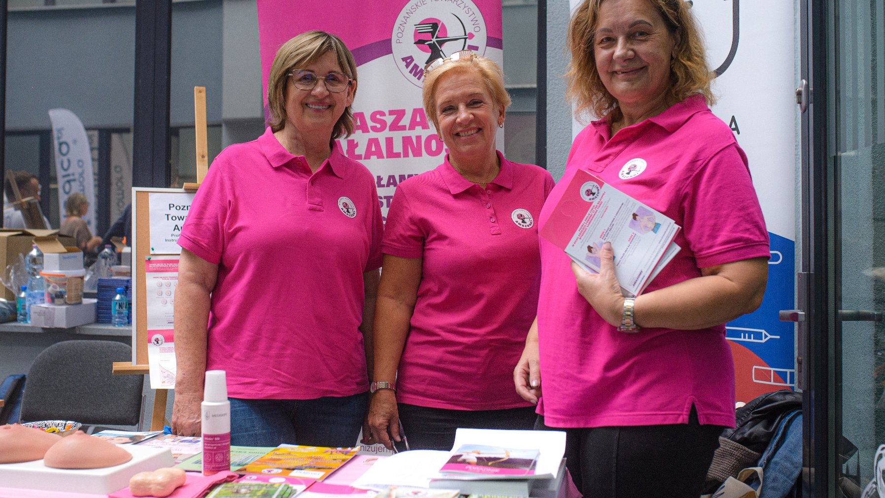 Na zdjęciu trzy kobiety w różowych koszulkach uśmiechają się do obiektywu