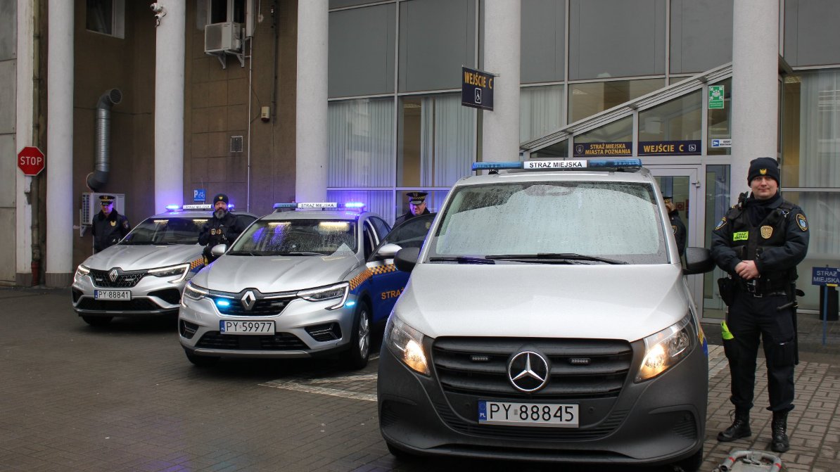 Na obrazie widać trzy pojazdy Straży Miejskiej zaparkowane przed budynkiem z dużymi oknami i kolumnami. Pojazdy to dwa samochody Renault van Mercedes z numerem rejestracyjnym PY 88845. Pojazdy mają niebieskie światła na dachu oraz napis "STRAŻ MIEJSKA", co wskazuje na to, że należą do miejskiej straży. Trzech strażników miejskich stoi w pobliżu pojazdów. Budynek posiada oznaczenia "WEJŚCIE C" i "STRAŻ MIEJSKA MIASTA POZNANIA", co sugeruje, że jest to wejście do straży miejskiej w Poznaniu.