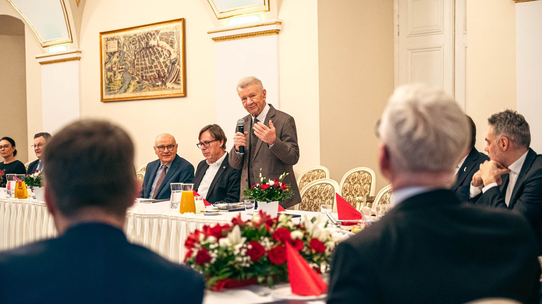 Formalne spotkanie w eleganckim pomieszczeniu z łukowymi sufitami i żyrondami. Wokół stołu pokrytego białym fartuchem siedzą osoby, a na stole znajdują się czerwone ręczniki, kwiaty i dzbanki z sokiem lub wodą. Jedna osoba stoi i wydaje się prowadzić prezentację lub mówić do grupy. W pokoju są także popiersie i ujęte w ramy dzieła sztuki na ścianach.