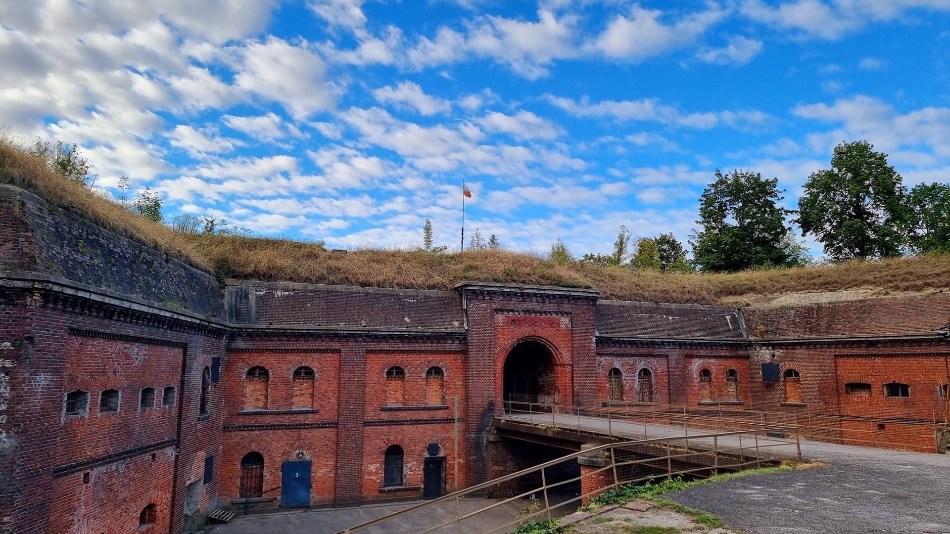 Historyczny ceglany fort z centralnym łukowatym wejściem. Budowla jest wbudowana w porośnięty trawą nasyp, na którym rośnie roślinność. Na szczycie nasypu znajduje się maszt z flagą. Fort ma kilka małych okien i drzwi, z niektórymi z nich okratowanymi. Metalowy mostek z poręczami prowadzi do głównego wejścia. Na niebie są niebieskie chmury.