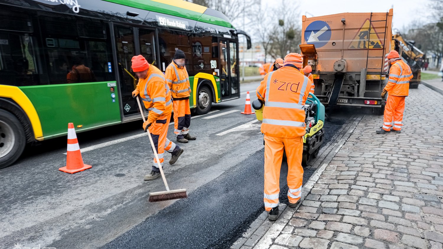 Ekipa pracowników Zakładu Robót Drogowych naprawia jezdnię. Obok widać przejeżdżający autobus