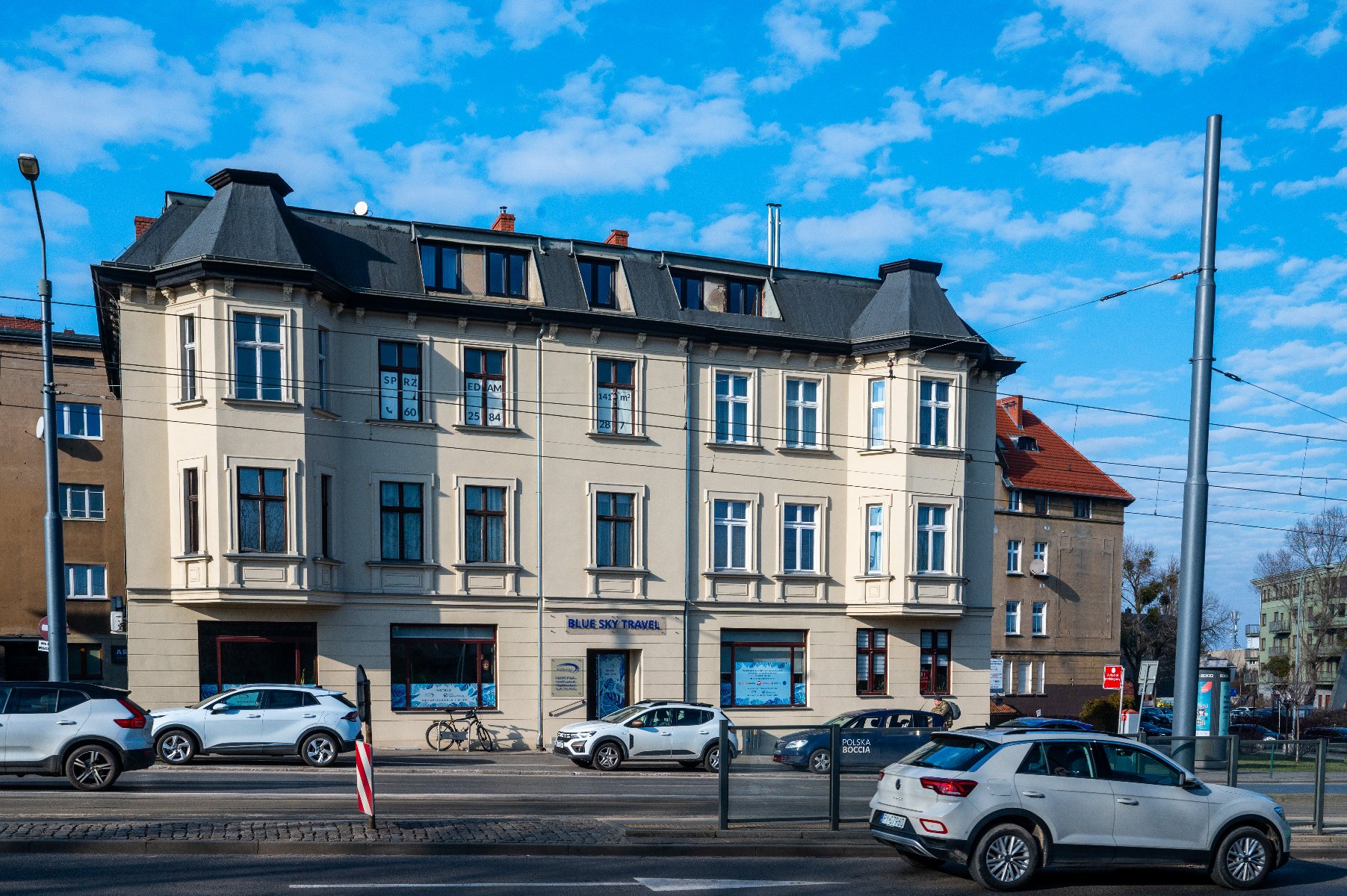 Na zdjęciu przedstawiony jest widok ulicy z budynkiem o beżowej fasadzie i ciemnym dachu. Budynek ma trzy piętra z wieloma oknami, z których niektóre mają znaki. Przed budynkiem, na ulicy, zaparkowane są samochody. Na parterze znajduje się firma o nazwie "BLUE SKY TRAVEL" z szyldem nad wejściem. Na niebie widać niebieskie niebo z rozproszonymi chmurami. - grafika artykułu