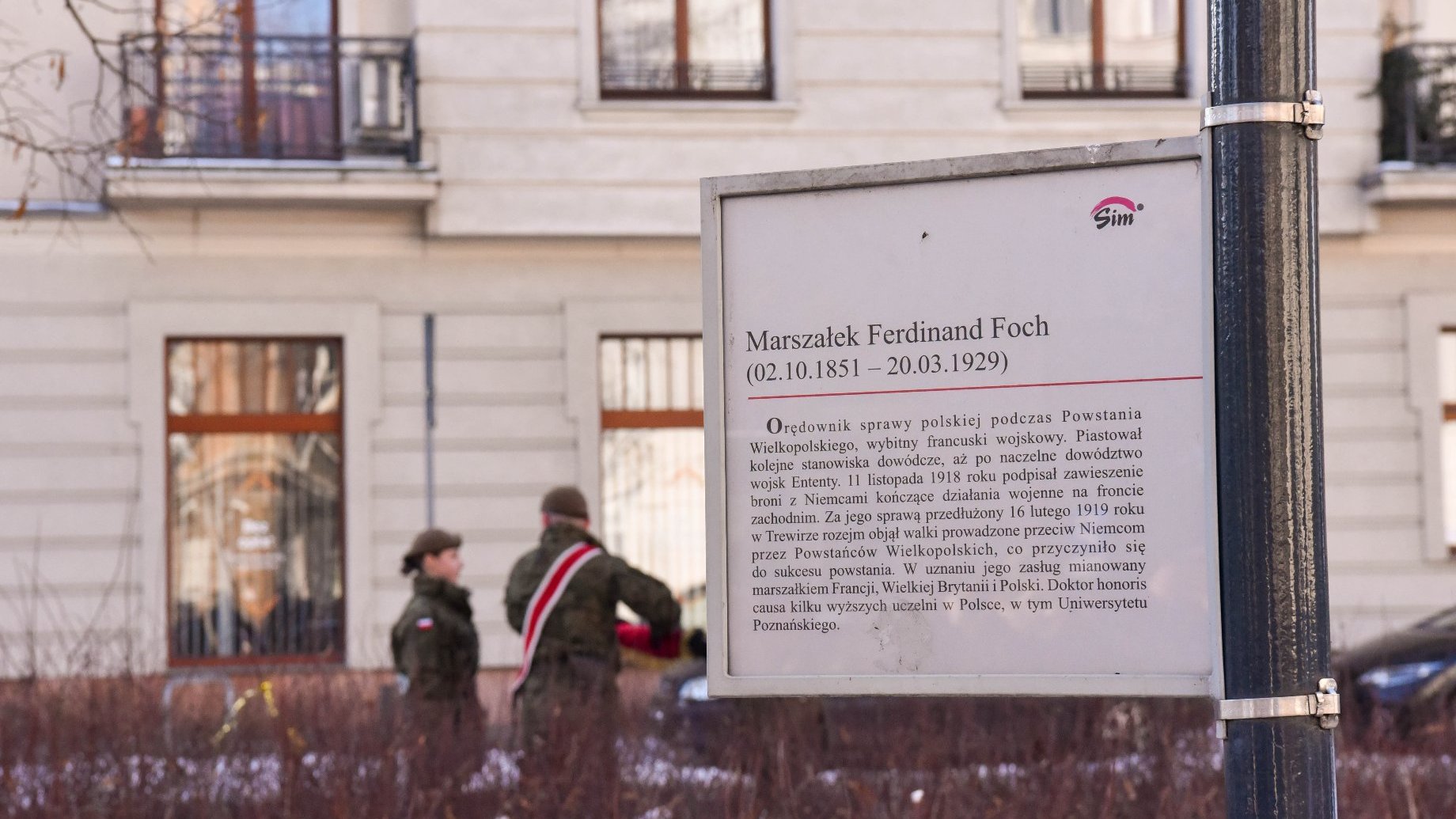 Tabliczka z dedykacją dla Marszałka Ferdinanda Focha, opisująca jego zasługi i znaczenie. Tabliczka jest w języku polskim i zawiera tekst: "Marszałek Ferdinand Foch (02.10.1851 - 20.03.1929) Ordownik sprawy polskiej podczas Powstania Wielkopolskiego, wybitny francuski wojskowy... (ciąg dalszy)." W tle znajdują się dwie osoby w mundurach wojskowych, jedna z nich ma na ramieniu biało-czerwoną szarfę.