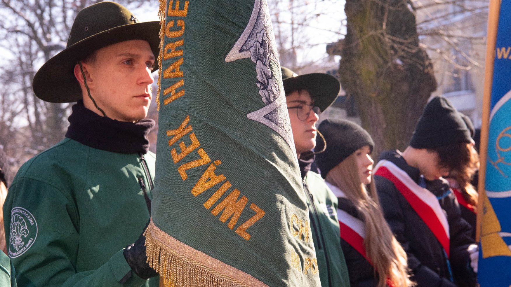 Grupa ludzi, z których część nosi zielone mundury i czapki, a reszta jest ubrana na czarno z czerwono-białymi szarfami. Jedna osoba na pierwszym planie trzyma zieloną flagę z złotymi frędzlami i tekstem 'ZWIĄZEK HARCERSKI'. Flaga ma również biały emblemat z kwiatowym wzorem.