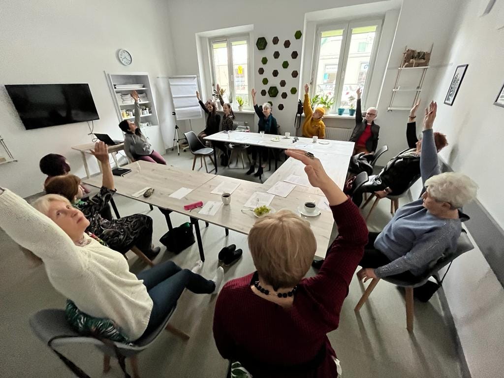 Na zdjęciu grupa ludzi przy stole, każdy z nich ma podniesioną w górę rękę - grafika artykułu