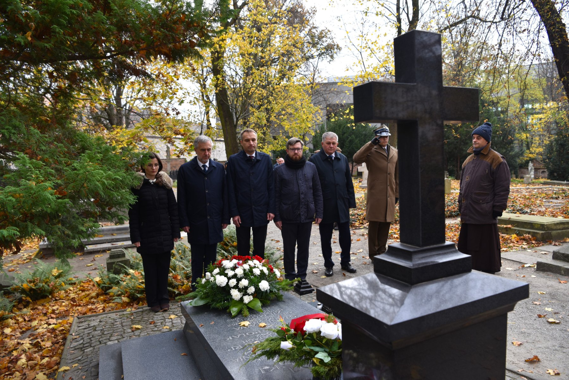 Grupa osób na cmentarzu, składają kwiaty na grobie, na ziemi leżą pożółkłe liście - grafika artykułu