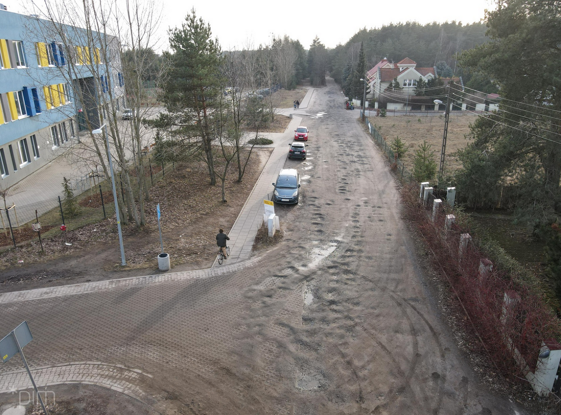 widok z lotu plata na ulicę nieutwardzoną, z jednej strony budynek, w tle drzewa, inny budynek, na ulic zaparkowane trzy samochody - grafika artykułu