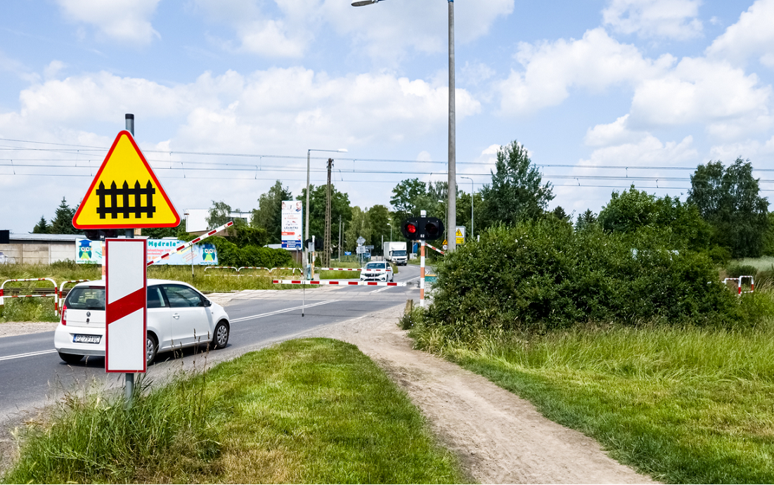 Przejazd kolejowy z zaporami widać samchód jadący w kierunku przejazdu, zapory są zamknięte - grafika artykułu