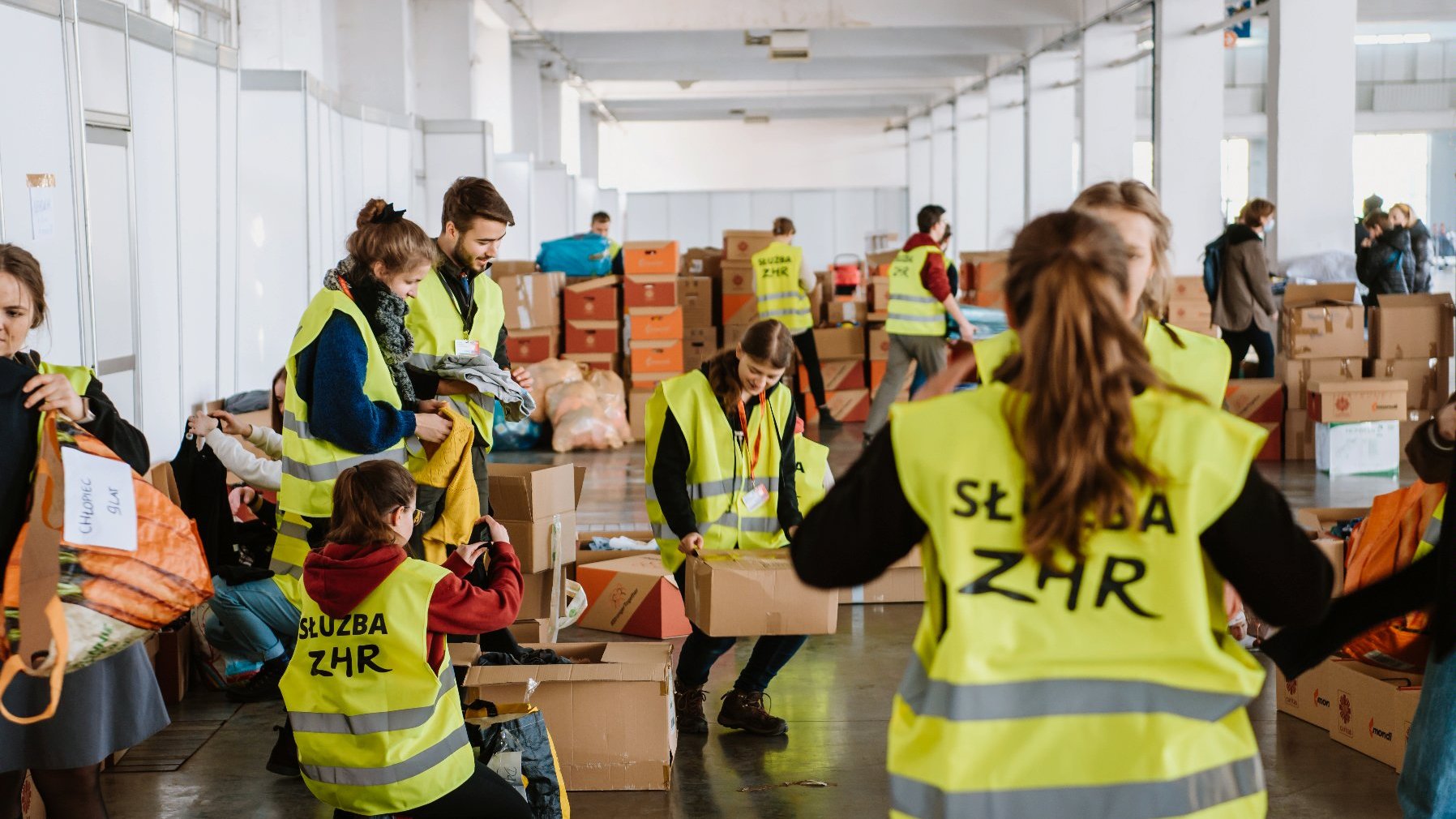 Zdjęcie przedstawia grupę osób w odblaskowych kamizelkach, które przenoszą paczki z pomocą humanitarną. - grafika artykułu