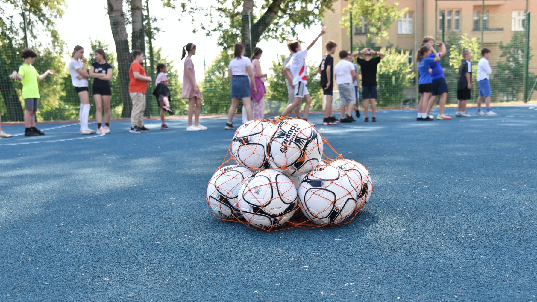 Zdjęcie przedstawia piłki w worku na boisku. W tle widać dzieci.