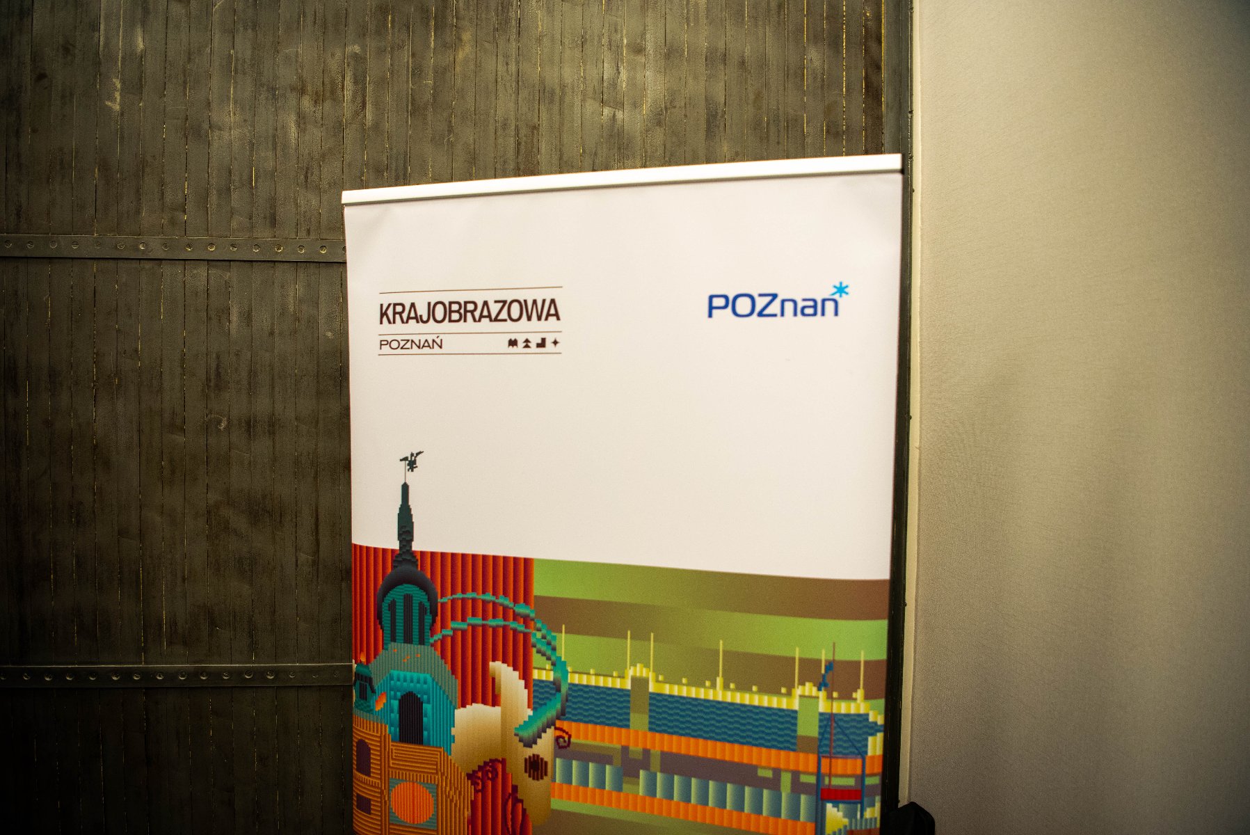 Plakat z napisem "KRAJOBRAZOWA POZNAŃ" i "POZnań". Plakat przedstawia kolorową, stylizowaną ilustrację elementów architektonicznych, prawdopodobnie reprezentujących zabytki lub budynki Poznania w Polsce. Tło plakatu jest białe, a ilustracja zawiera różne kolory, takie jak czerwony, zielony, niebieski i żółty. Plakat jest umieszczony na tle teksturowanej, ciemnej ściany po lewej i gładkiej, jasnej ściany po prawej stronie. - grafika artykułu