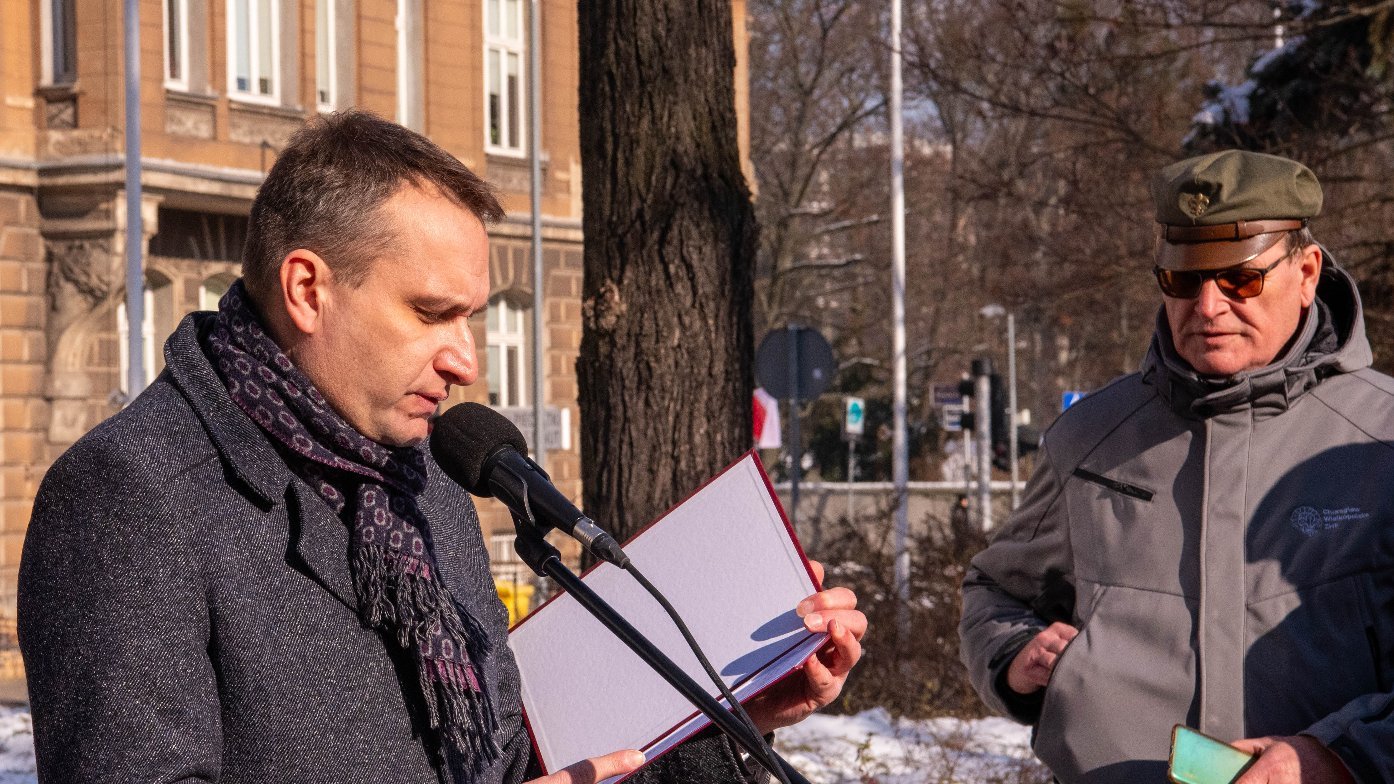 Dwóch mężczyzn na zewnątrz, jeden mówi do mikrofonu, trzymając otwartą książkę lub folder, drugi stoi z boku, trzymając smartfon. W tle budynek i śnieg na ziemi.