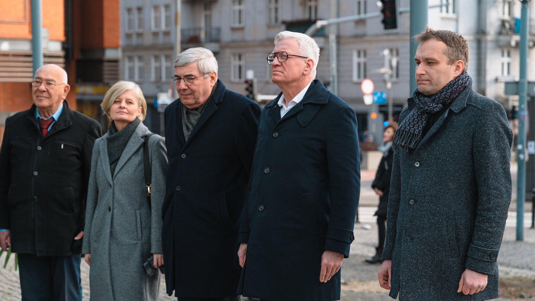 Pięć osób stojących w miejskiej scenerii na zewnątrz. Ubrane są formalnie lub pół-formalnie, w tym w płaszcze i szaliki, co sugeruje chłodną pogodę. W tle widoczne są budynki, sygnalizacja świetlna i znaki drogowe, co wskazuje, że jest to prawdopodobnie wydarzenie publiczne lub oficjalne. Ludzie stoją w rzędzie, prawdopodobnie uczestnicząc lub obserwując ceremonię lub zgromadzenie.