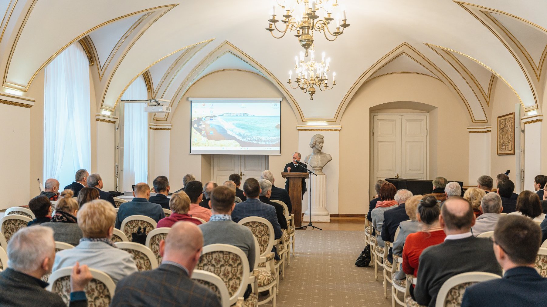 Formalne wydarzenie w eleganckiej sali z łukowatymi sufitami i żyrandolami. Ludzie siedzą na ozdobnych krzesłach, skierowanych w stronę mówcy stojącego przy podium. Za mówcą duży ekran wyświetlający nadmorską scenerię. W tle popiersie i fortepian.