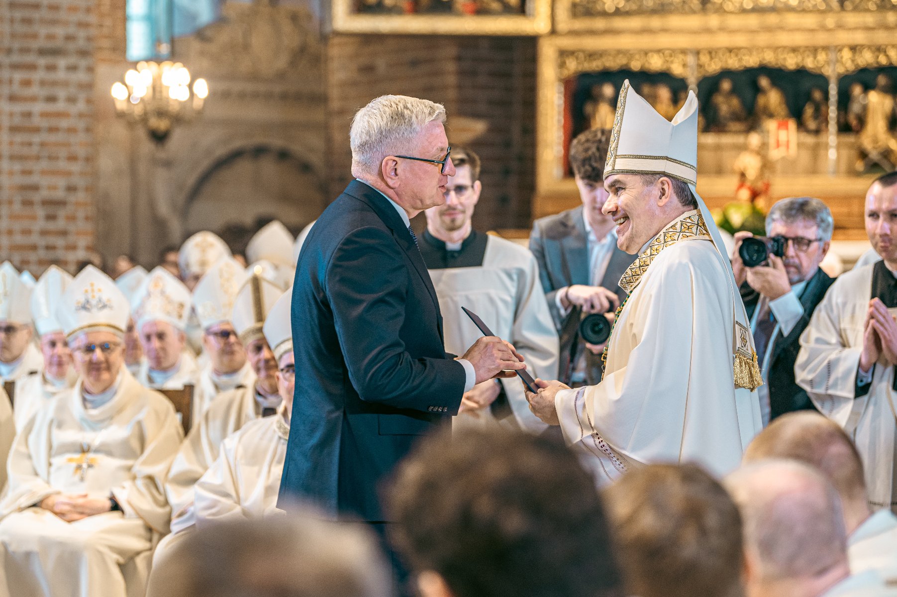 1 maja odbył się ingres arcybiskupa Zbigniewa Zielińskiego Wnętrze kościoła podczas ważnej uroczystości religijnej. Po prawej stronie mężczyzna w ciemnym garniturze odbiera dokument od duchownego ubranego w biało-złote szaty liturgiczne. W tle widoczni są inni duchowni w białych strojach, siedzący i obserwujący wydarzenie. Na suficie ozdobne detale, w tym żyrandol.