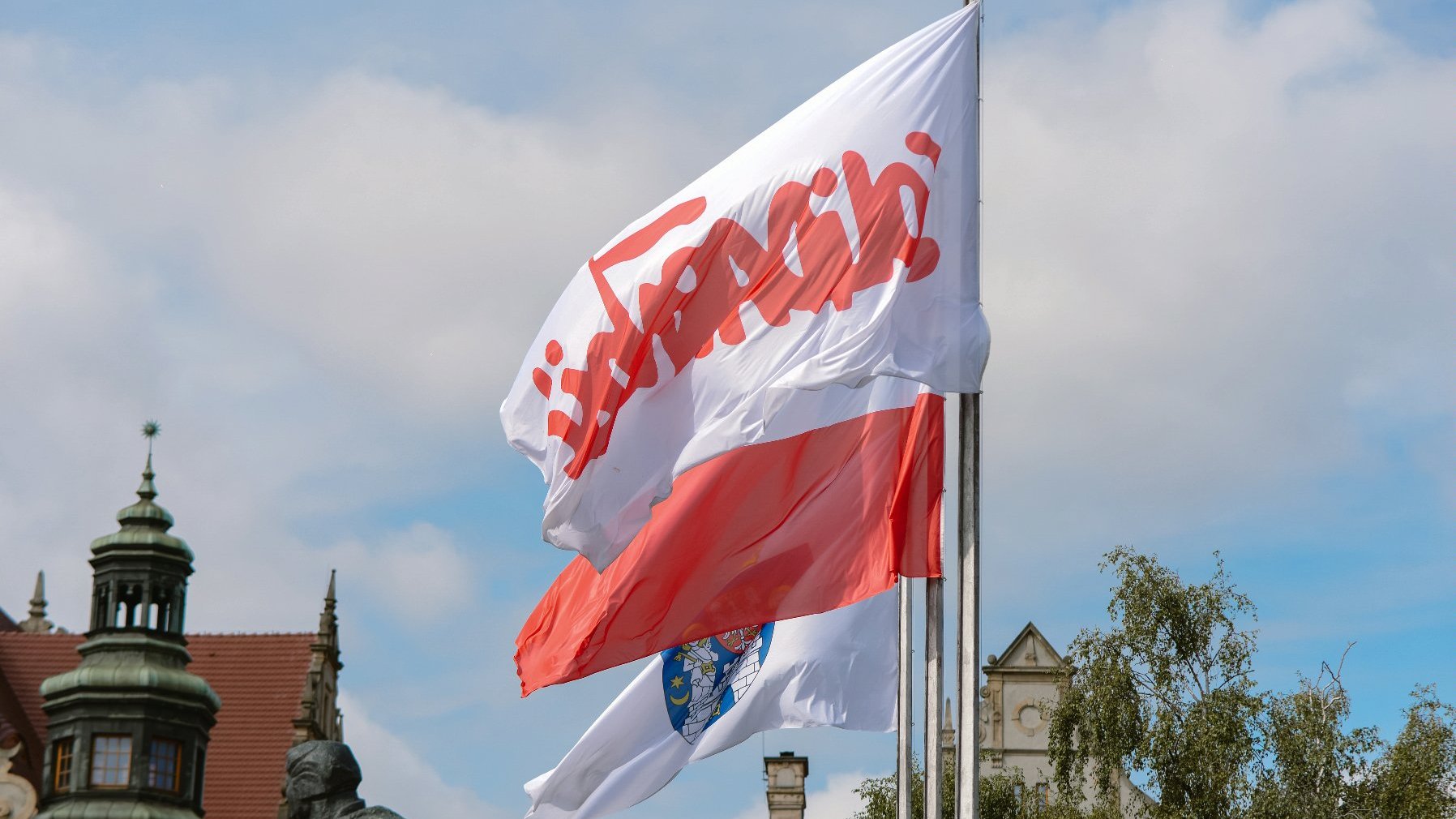 Zdjęcie przedstawia flagi Polski, z herbem Poznania i znakiem Solidarności. W tle znajduje się pomnik Dwóch Krzyży.