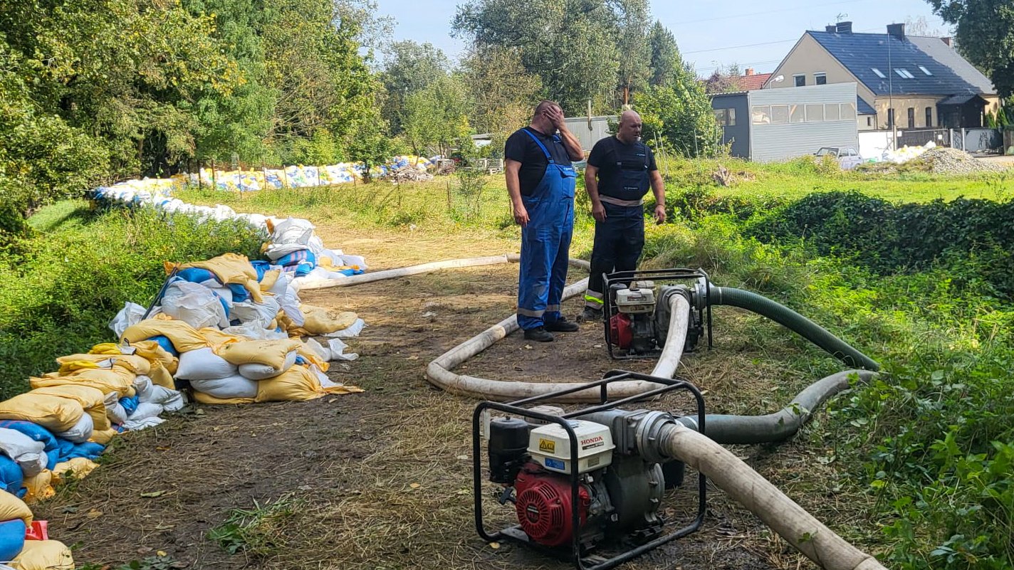 Zdjęcie przedstawia dwóch mężczyzn podczas pracy. Obok leżą worki z piaskiem.