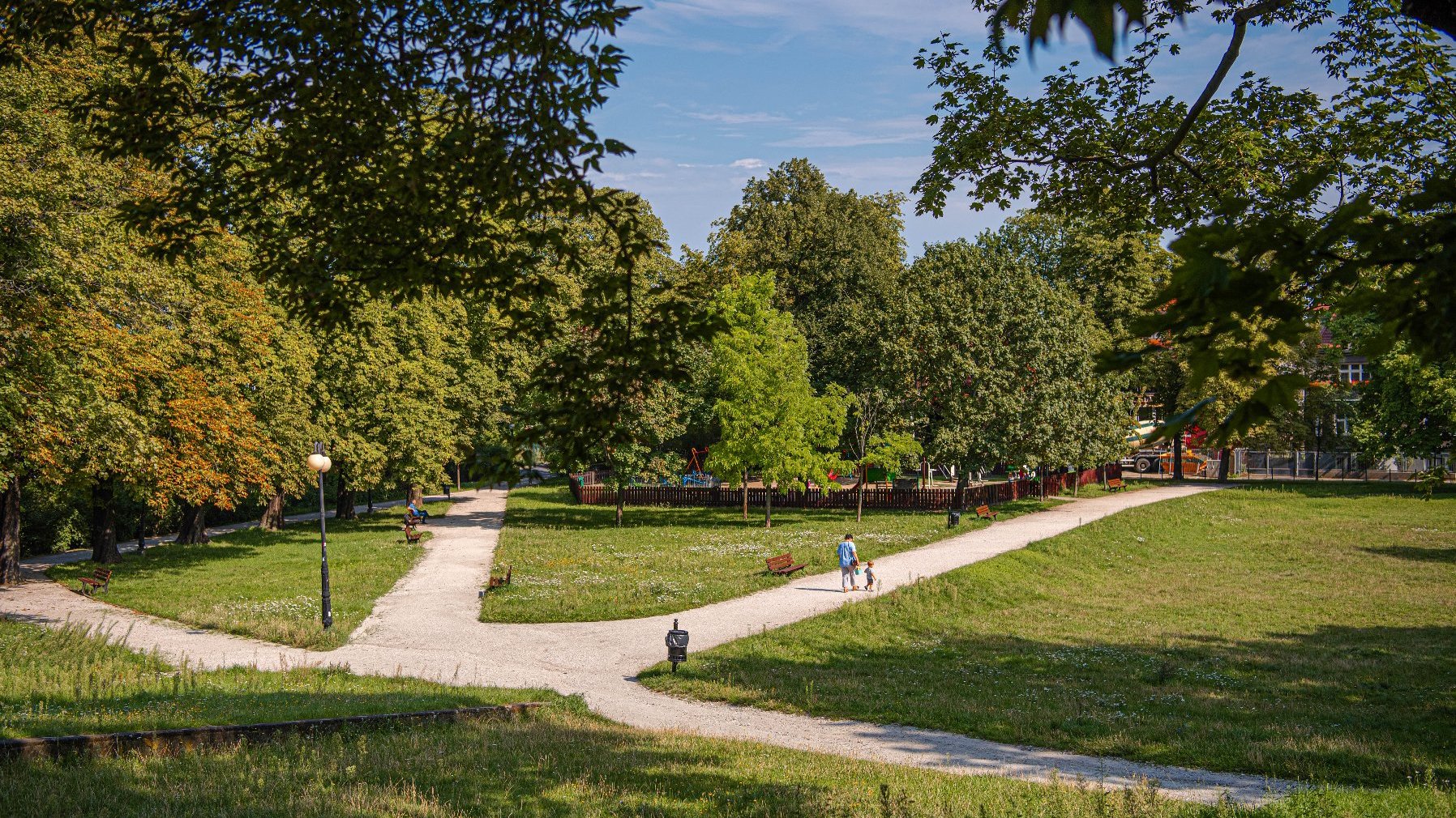 Zdjęcie przedstawia park. Widać na nim zieleń - trawę i drzewa oraz alejki z ławkami, a także spacerujących ludzi. W tle znajduje się ogrodzenie, za którym jest plac zabaw.