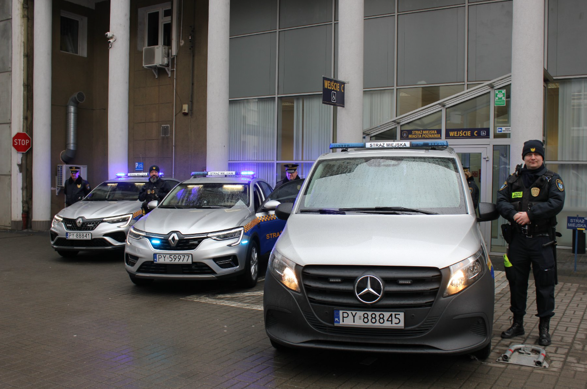 Na obrazie widać trzy pojazdy Straży Miejskiej zaparkowane przed budynkiem z dużymi oknami i kolumnami. Pojazdy to dwa samochody Renault van Mercedes z numerem rejestracyjnym PY 88845. Pojazdy mają niebieskie światła na dachu oraz napis "STRAŻ MIEJSKA", co wskazuje na to, że należą do miejskiej straży. Trzech strażników miejskich stoi w pobliżu pojazdów. Budynek posiada oznaczenia "WEJŚCIE C" i "STRAŻ MIEJSKA MIASTA POZNANIA", co sugeruje, że jest to wejście do straży miejskiej w Poznaniu. - grafika artykułu