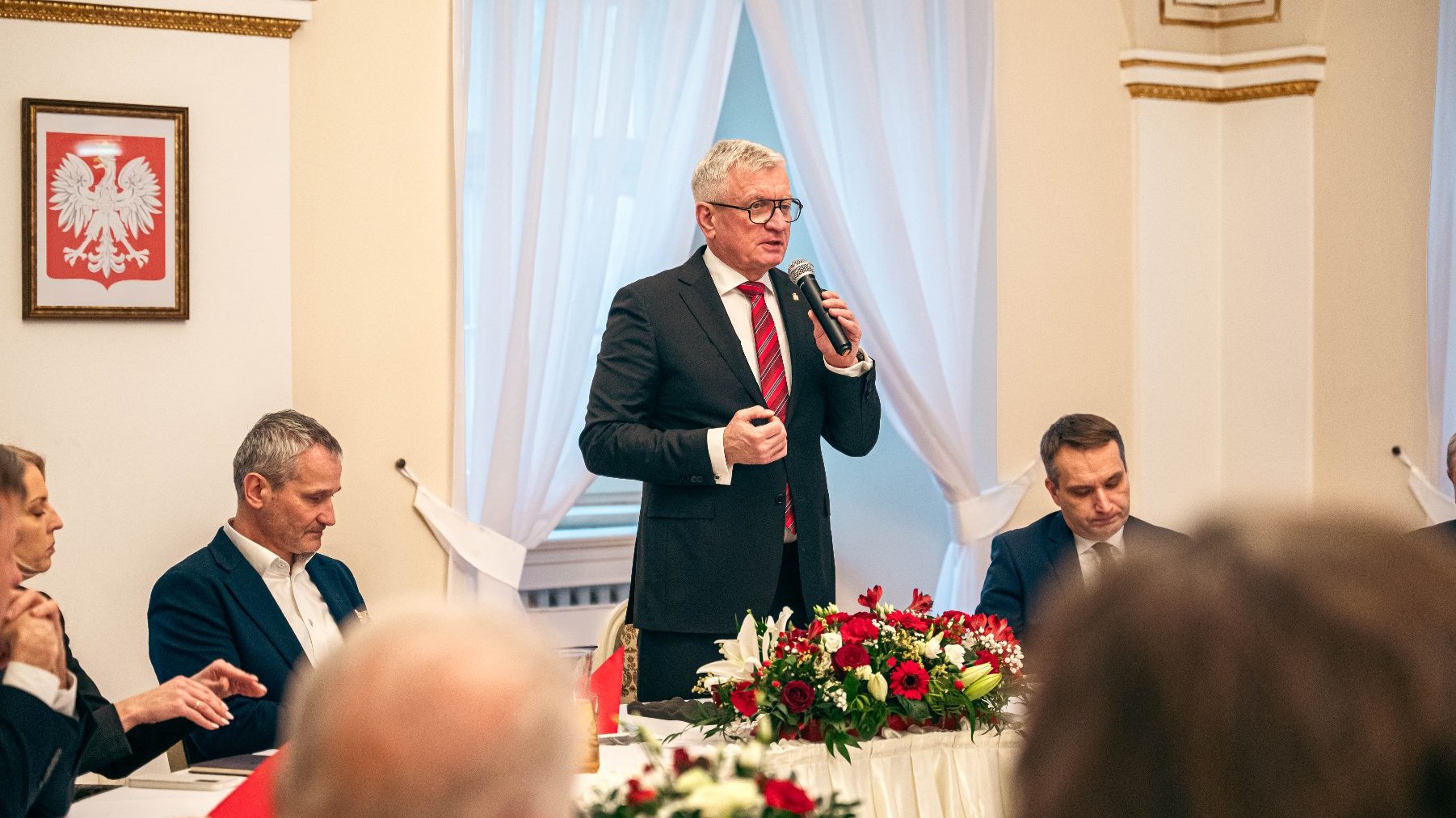 Zdjęcie przedstawia formalne spotkanie lub konferencję, na której siedzą kilka osób wokół stołu. Centralna postać stoi i mówi przez mikrofon, ubrana w garnitur i krawat w czerwone i białe paski. Stołek ozdobiony jest kwiatami, a na ścianie znajduje się wizerunek białego orła na czerwonym tle, który jest często kojarzony z Polską.