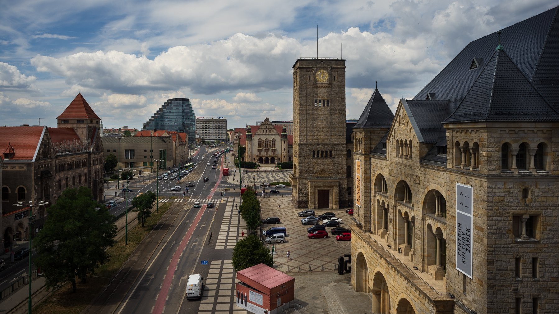 Scena miejska z mieszanką historycznej i nowoczesnej architektury. Na pierwszym planie widoczny jest duży kamienny budynek z łukowatymi oknami i wieżą zegarową. Przed budynkiem zaparkowanych jest kilka samochodów. Po lewej stronie znajduje się kolejny historyczny budynek z czerwonym dachówką. W tle wyróżnia się nowoczesny, unikatowy budynek na tle innych, bardziej konwencjonalnych struktur. Ulica pośrodku ma torowisko tramwajowe, ścieżkę rowerową i kilka pojazdów, w tym biały van.