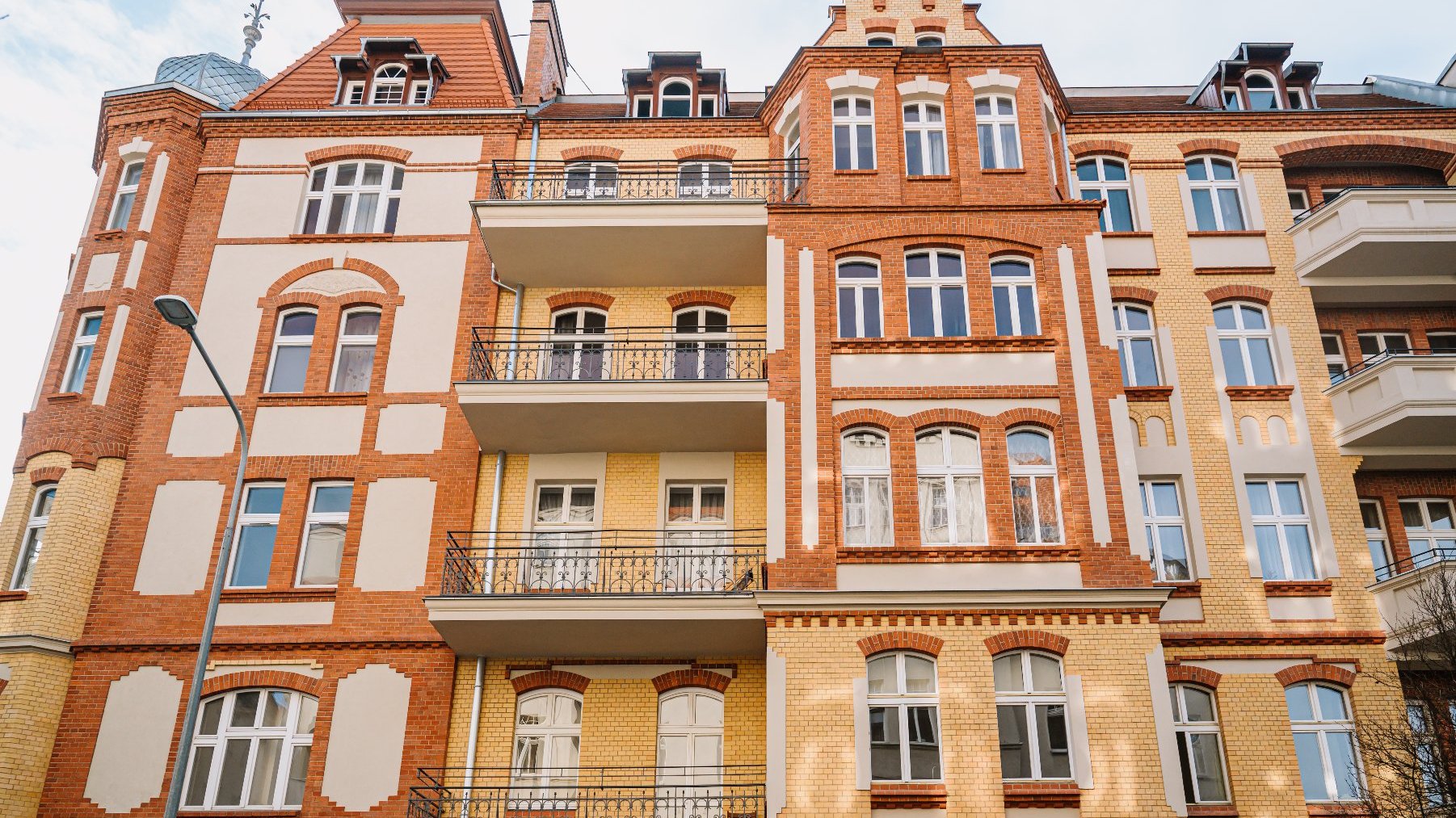 Fasada wielopiętrowego budynku mieszkalnego o zróżnicowanym stylu architektonicznym. Budynek łączy cegłę czerwoną z jasnym tynkiem, z ozdobnymi elementami wokół łukowych okien oraz balkonów z kutymi żelaznymi balustradami. Dach ma okna połaciowe oraz małą iglicę. Cenna wizualnie kombinacja historycznych i nowoczesnych elementów architektonicznych.