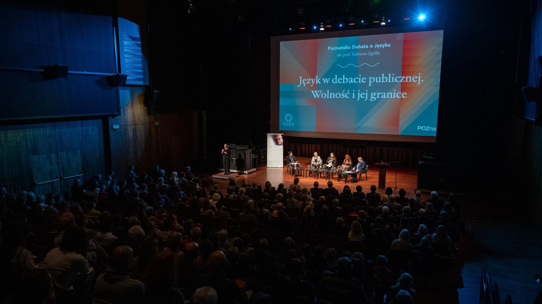 Na zdjęciu grupa ludzi zebranych w sali CK Zamek, na scenie widać uczestników debaty, za nimi ekran