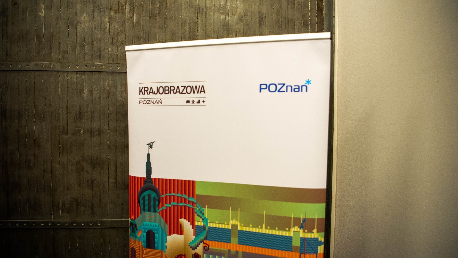 Plakat z napisem "KRAJOBRAZOWA POZNAŃ" i "POZnań". Plakat przedstawia kolorową, stylizowaną ilustrację elementów architektonicznych, prawdopodobnie reprezentujących zabytki lub budynki Poznania w Polsce. Tło plakatu jest białe, a ilustracja zawiera różne kolory, takie jak czerwony, zielony, niebieski i żółty. Plakat jest umieszczony na tle teksturowanej, ciemnej ściany po lewej i gładkiej, jasnej ściany po prawej stronie.