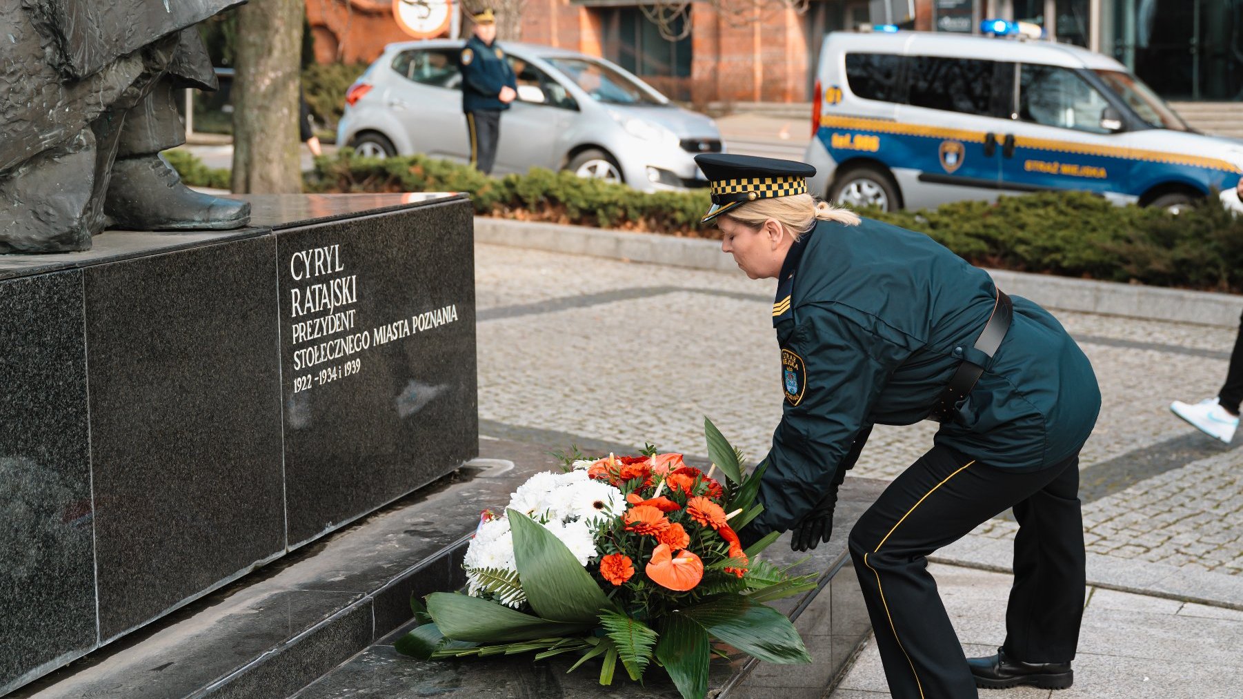 Osoba w mundurze, prawdopodobnie policjant lub pracownik ochrony, umieszcza wieniec kwiatowy u podstawy pomnika. Na pomniku widnieje napis: "CYRYL RATAJSKI PREZYDENT STOŁECZNEGO MIASTA POZNANIA 1922-1924 I 1934-1939" W tle znajdują się dwa pojazdy, z których jeden to pojazd policyjny, oraz inna osoba w mundurze stojąca w pobliżu pojazdów.