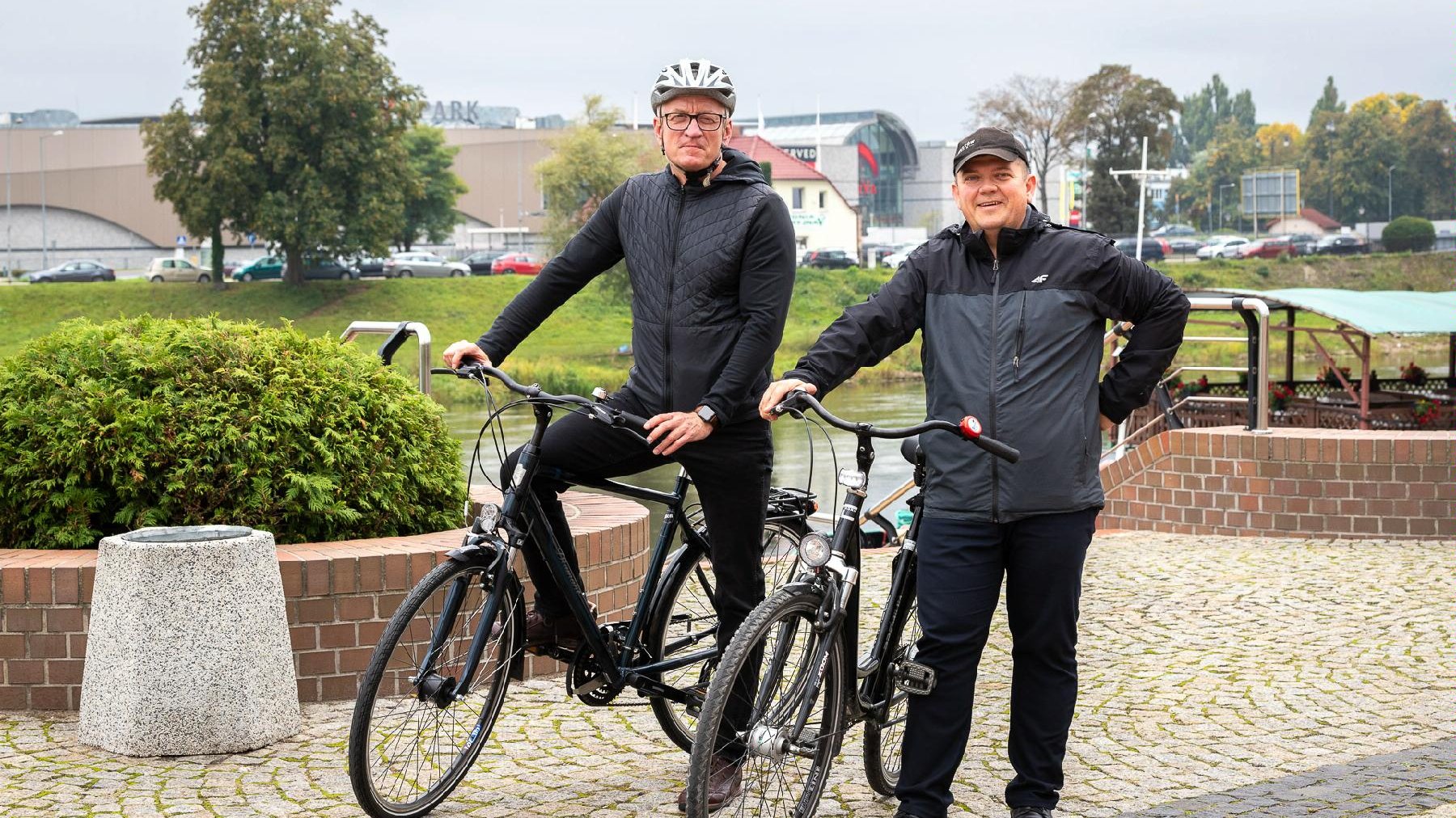 Gorzów, bulwar. Prezydenci Jacek Jaśkowiak i Jacek Wójcicki stoją obok siebie, prezydent Jaśkowiak na rowerze, prezydent Wójcicki stoi obok roweru. Obaj patrzą w obiektyw. W tle rzeka i zabudowania