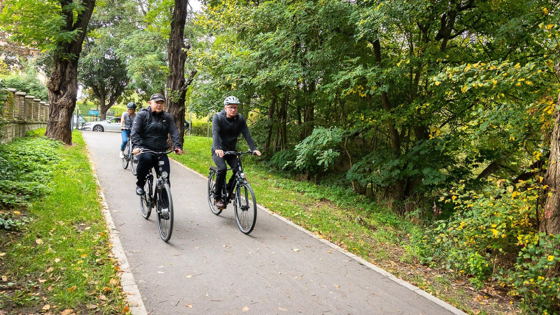 Prezydenci Jacek Jaśkowiak i Jacek Wójcicki na wspólnej przejażdżce rowerowej. Jadą drogą rowerową, po obu jej stronach drzewa i rośliny. Za nimi inny rowerzysta