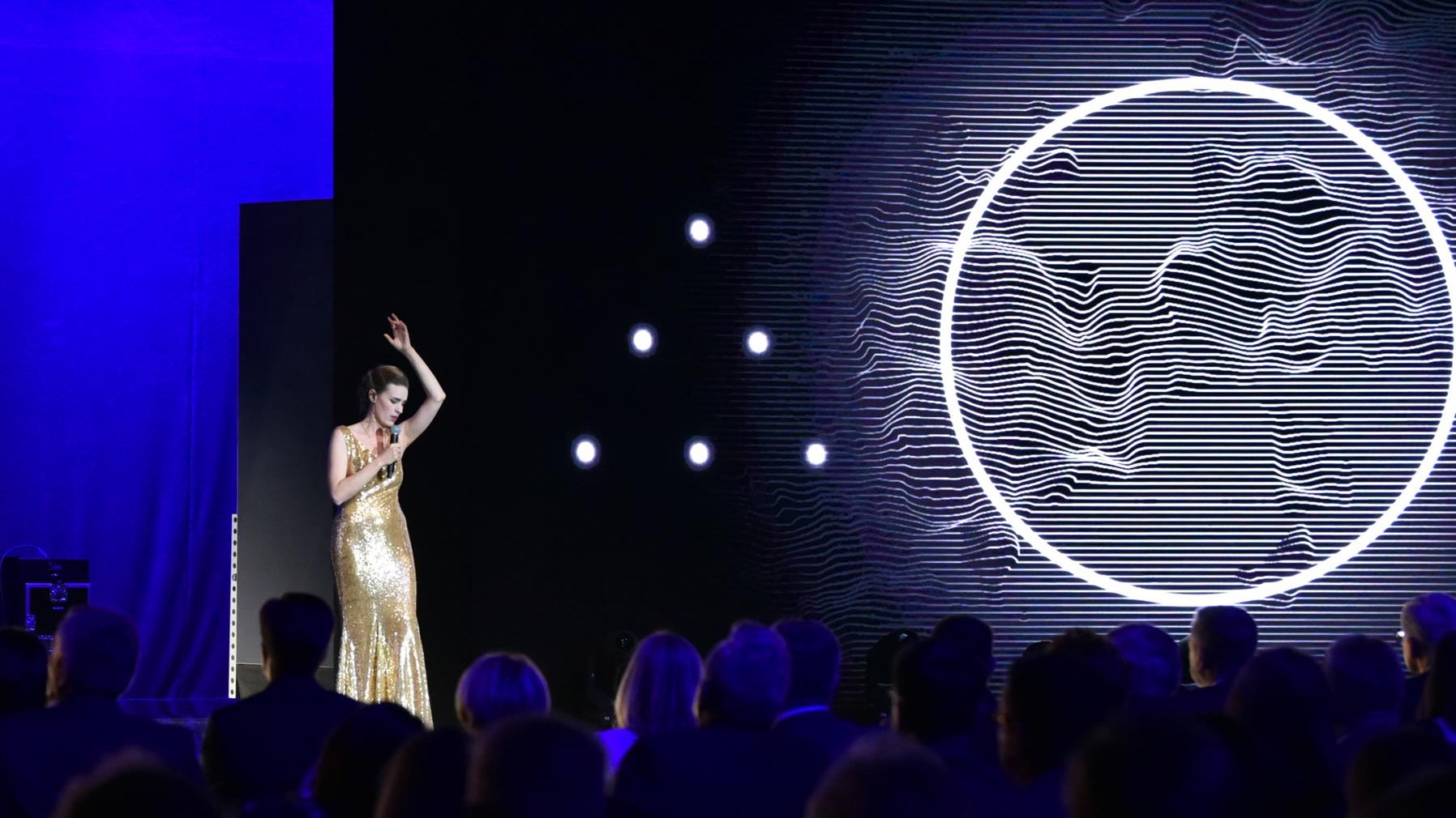 Na zdjęciu śpiewająca kobieta w złotej sukni, obok, na ekranie, widać wielkie koło