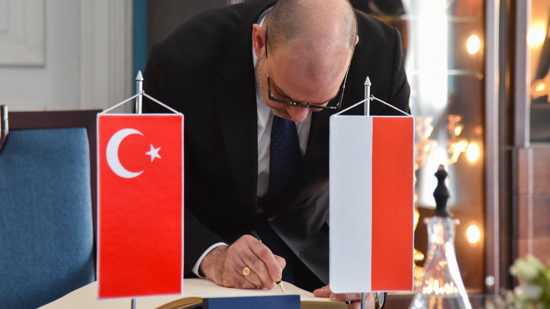 Na zdjeciu mężczyzna wpisujący się do księgi pamiątkowej, obok niej stoją dwie flagi: polska i turecka