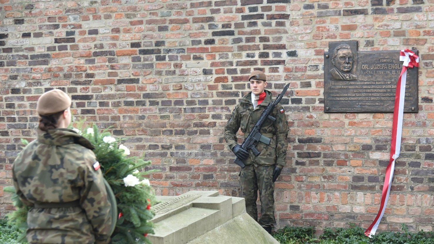 Obraz przedstawia ceremonię wojskową odbywającą się przed tablicą pamiątkową umieszczoną na ceglanej ścianie. Obecnych jest trzech żołnierzy w mundurach kamuflażowych. Jeden żołnierz trzyma wieniec, podczas gdy pozostali dwaj stoją na straży z karabinami. Tablica pamiątkowa jest ozdobiona czerwoną i białą wstążką. Przed tablicą znajduje się kamienny pomnik otoczony zielenią.