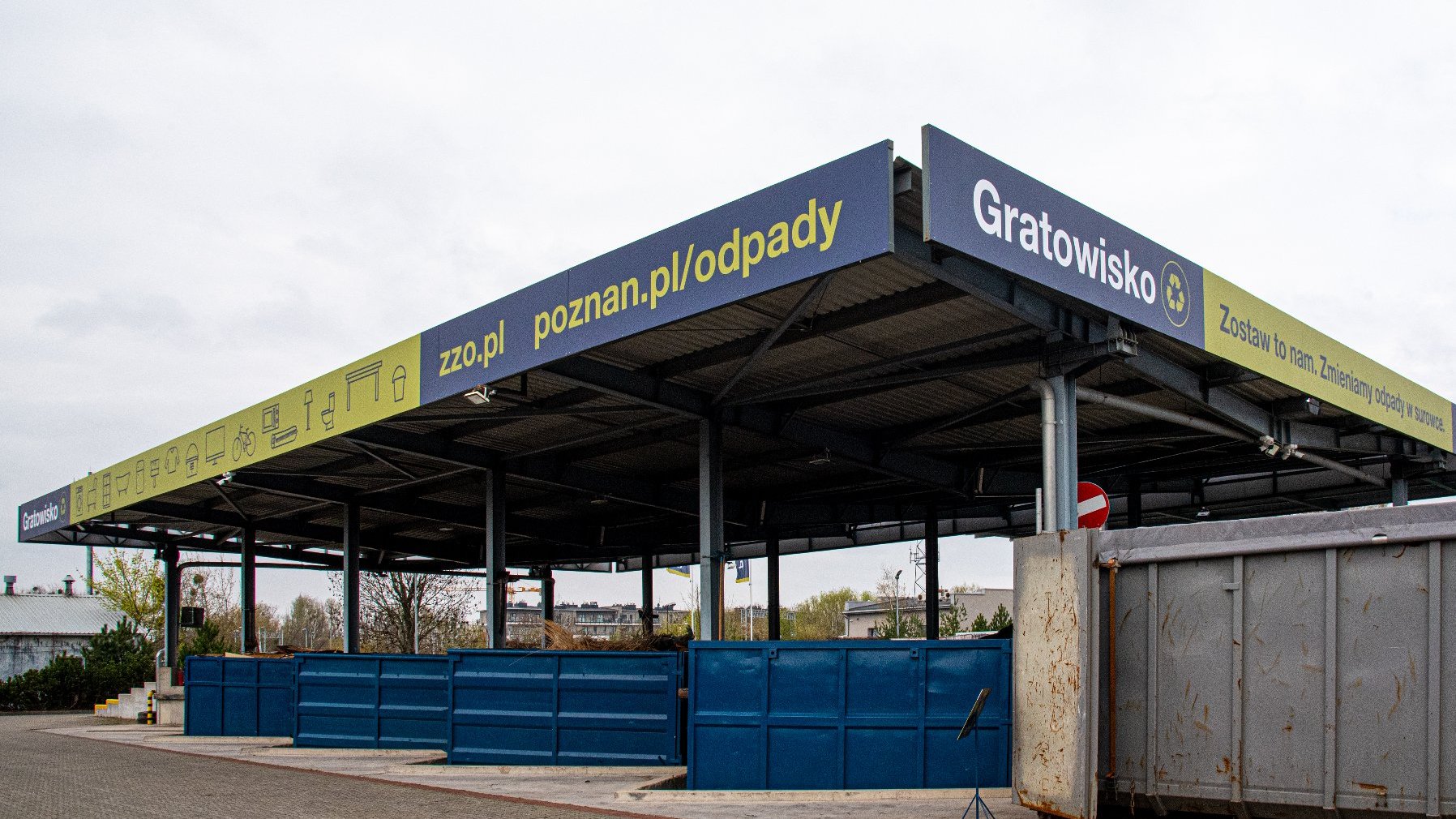 Obraz przedstawia punkt zbiórki odpadów lub centrum recyklingu. Konstrukcja to zadaszona przestrzeń z niebieskimi kontenerami lub pojemnikami do sortowania różnych typów odpadów. Na oznakowaniach na konstrukcji widnieją napisy 'Gratowisko' oraz adresy URL 'zzo.pl' i 'poznan.pl/odpady'. Znaki na konstrukcji zawierają także różne ikony reprezentujące różne typy odpadów, takie jak elektronika, meble i materiały niebezpieczne.