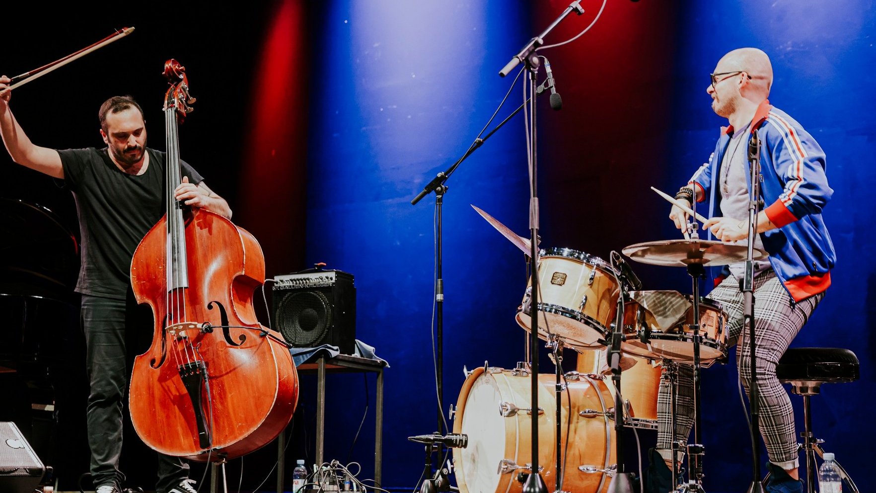 Koncert na scenie. Jeden z muzyków gra na kontrabasie za pomocą smyczka, drugi muzyk gra na perkusji. Scena oświetlona czerwonymi i niebieskimi światłami. Przed muzykami ustawione są mikrofony. Na scenie widać również wzmacniacz i kable. Tło jest ciemne, co podkreśla muzyków i ich instrumenty.