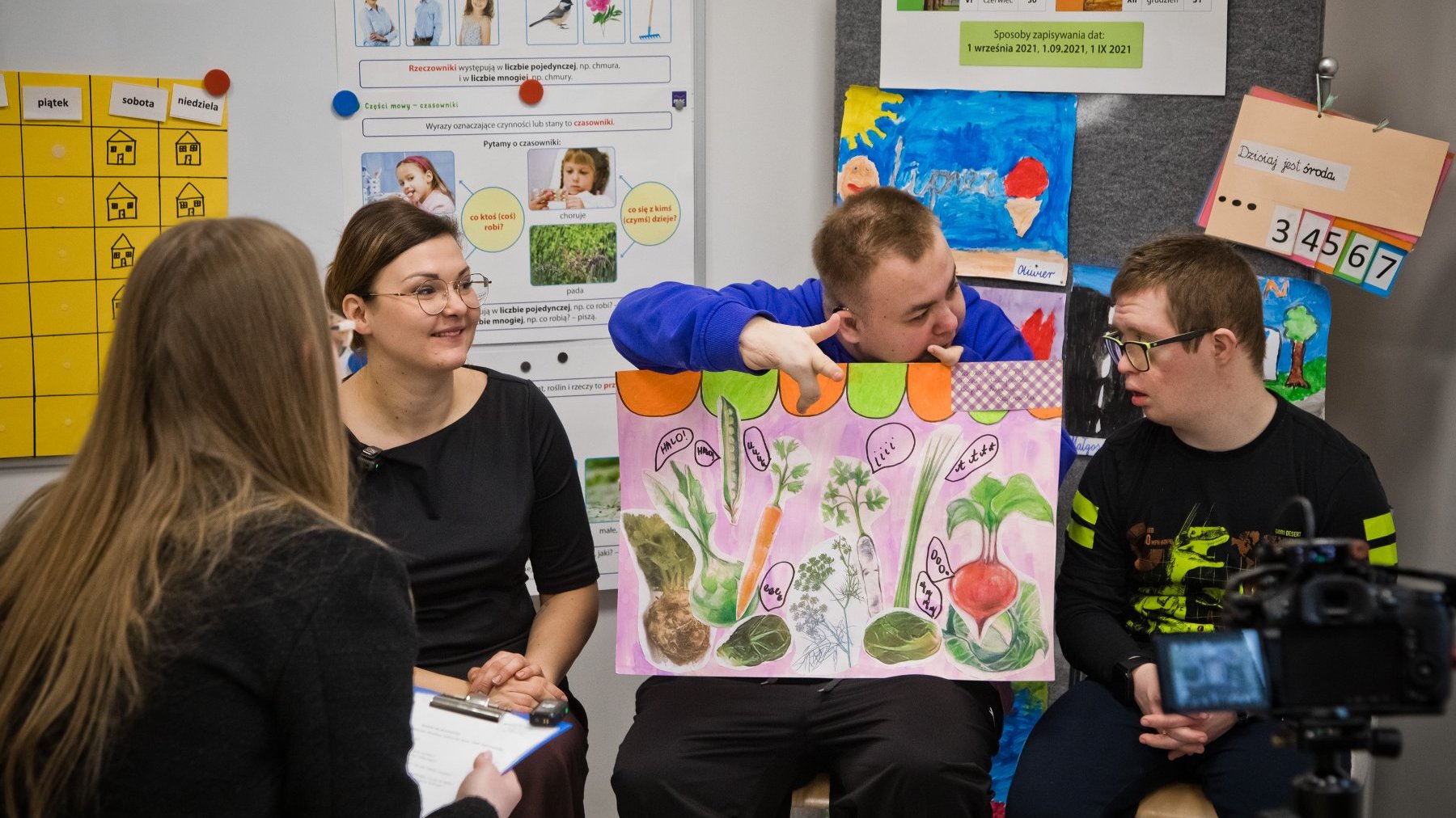 Zdjęcie grupy osób w przestrzeni edukacyjnej. Na ścianach widoczne różne materiały edukacyjne i plakaty, w tym kalendarz z dniami tygodnia po polsku ("piątek", "sobota", "niedziela") oraz ilustracje przedstawiające warzywa (m.in. marchewkę, rzodkiewkę) z ich nazwami. Jedna z osób trzyma duży, kolorowy plakat z rysunkami warzyw.