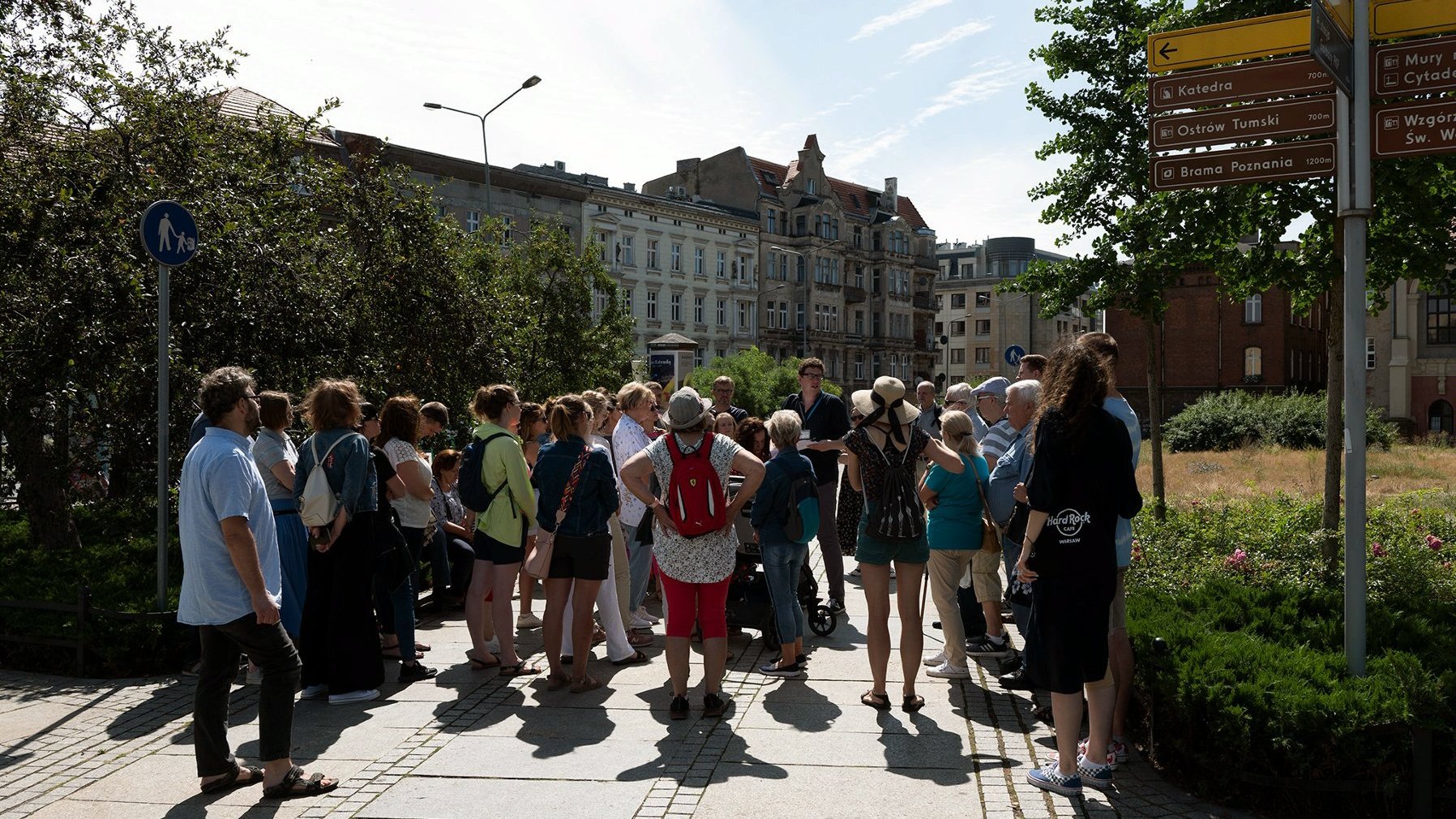 Grupa uczestników przechadzki stojąca na chodniku przed drogowskazem na Ostrów Tumski.
