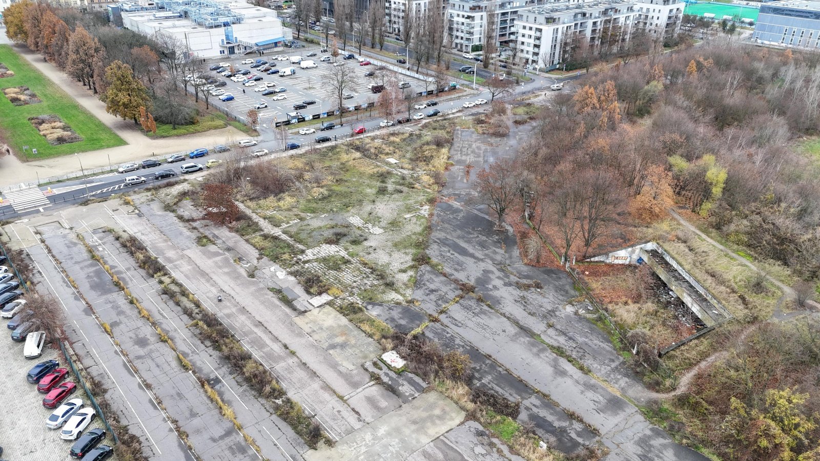 Widok z lotu ptaka na powierzchnię pokrytą niszczejącym betonem i starym asfaltem. W tle budynki osiedla mieszkaniowego.