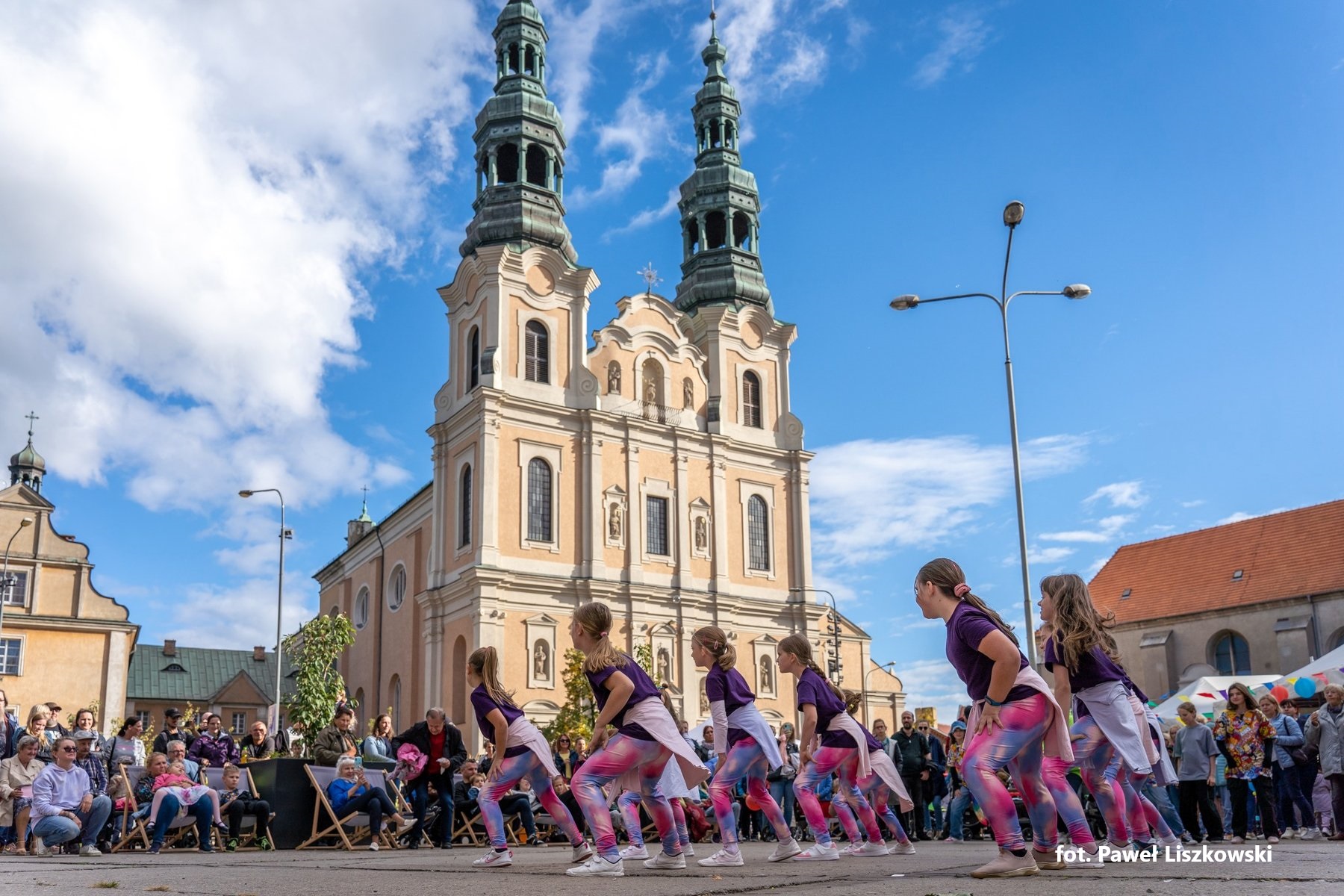 Na płycie Placu Bernardyńskiego tańczy przed publicznością zespół dziewczynek. W tle kościół i kamienice. - grafika artykułu