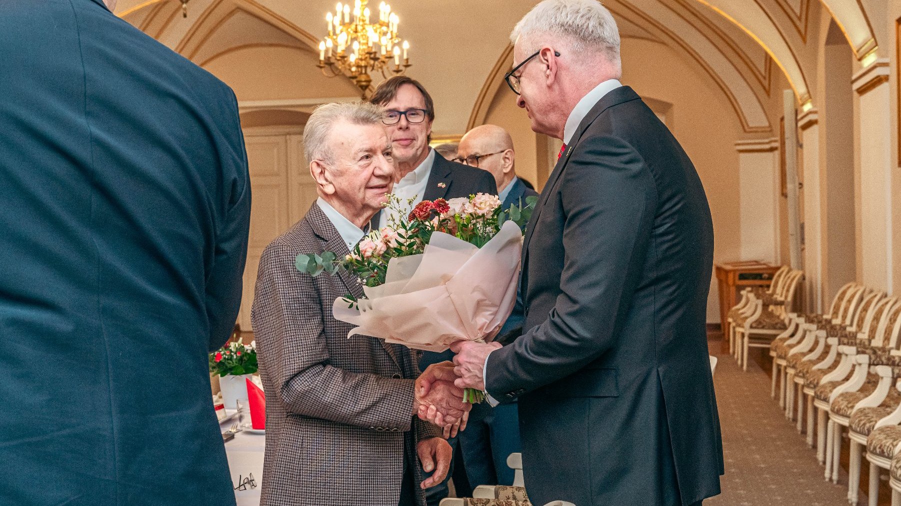 "Formalne spotkanie w eleganckim pomieszczeniu z łukowymi sufitami i żyrondami. Jedna osoba wręcza drugiej bukiet kwiatów.