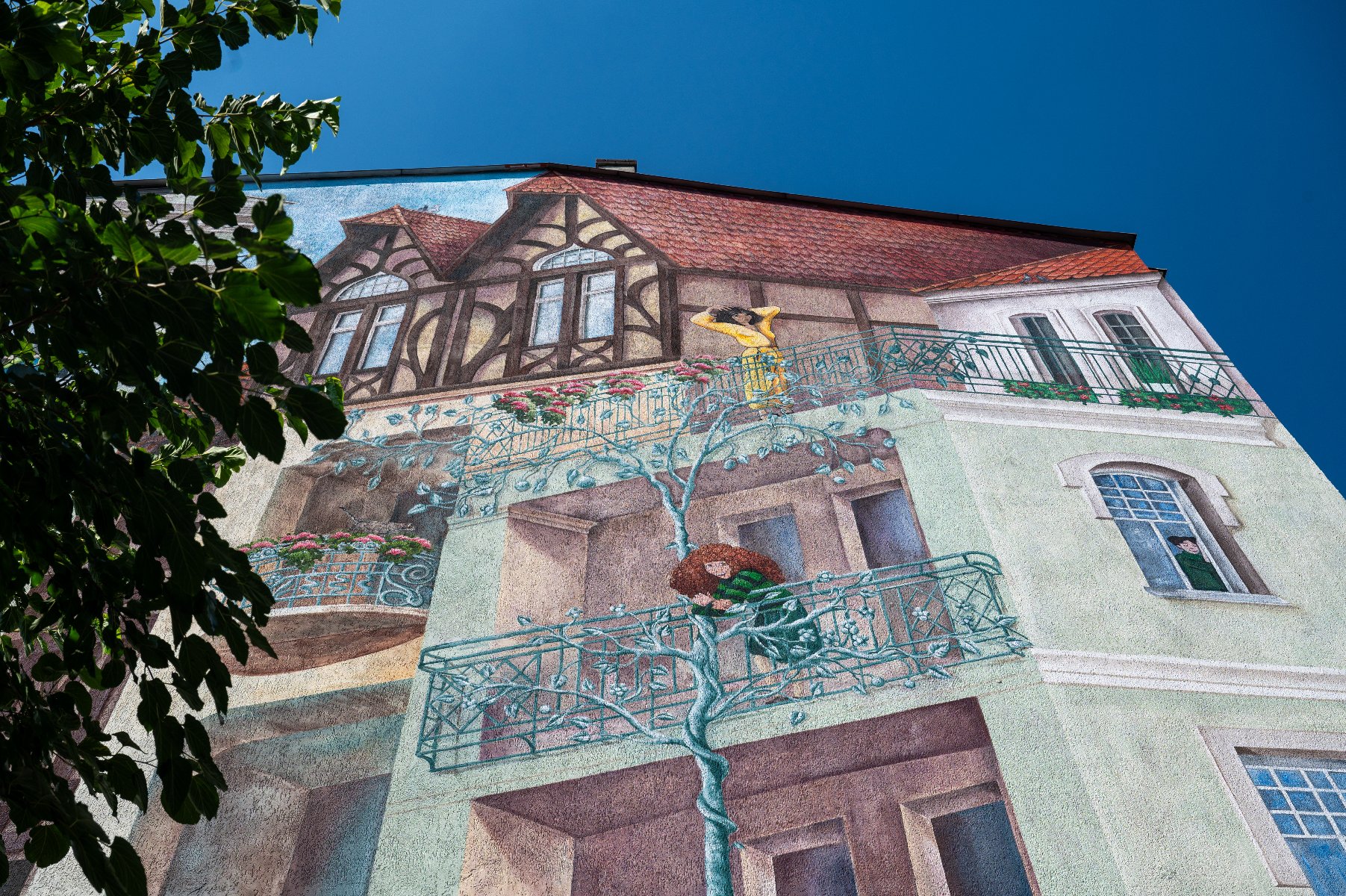 Malowidło ścienne na elewacji budynku, które przedstawia iluzję architektoniczną. Na malowidle widoczny szczegółowy obraz innego wielopiętrowego budynku z balkonami, oknami oraz drzewem wspinającym się po jego ścianie. Na balkonie stoi postać, z okna spogląda druga osoba, a na gałęzi namalowanego drzewa znajduje się ptasie gniazdo z ptakiem. Na górze malowidła znajduje się pół-drewniany dom z czerwonym dachem. - grafika artykułu