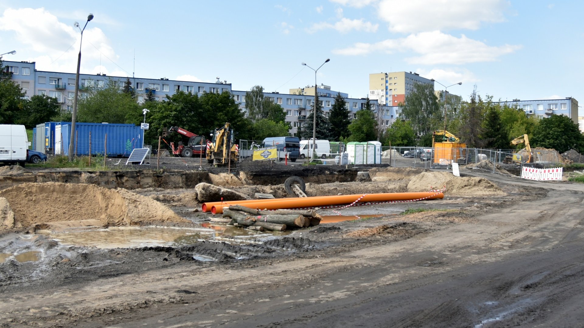 Zdjęcie przedstawia plac budowy, widać na nim elementy budowlane i hałdy ziemi. W tle znajdują się bloki