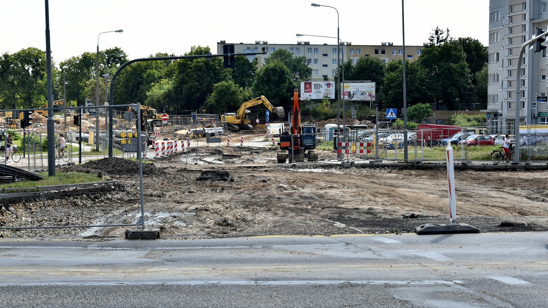 Na zdjęciu znajduje się plac budowy, pracują koparki, na dalszym planie widać bloki