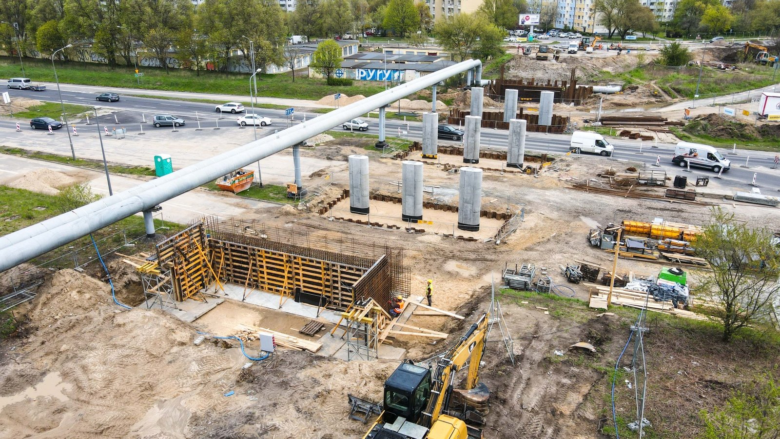 Budowa wiaduktu. Rury ciepłownicze i podpory wiaduktu. W głębi bloki i drzewa.